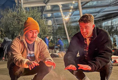 Two young males crouched around a fire at the Eden Project, wearing coats and hats, warming their hands up